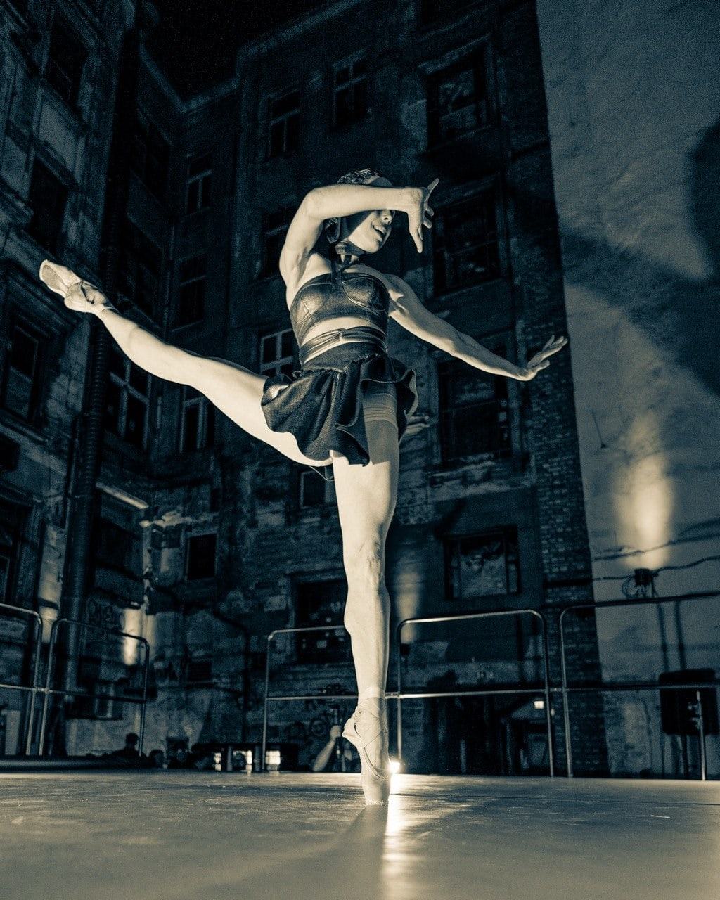 Ballett-Tänzerin in der Arabesque auf dem beleuchteten Bühnenboden im nächtlichen Hof, ein Arm vor dem Gesicht